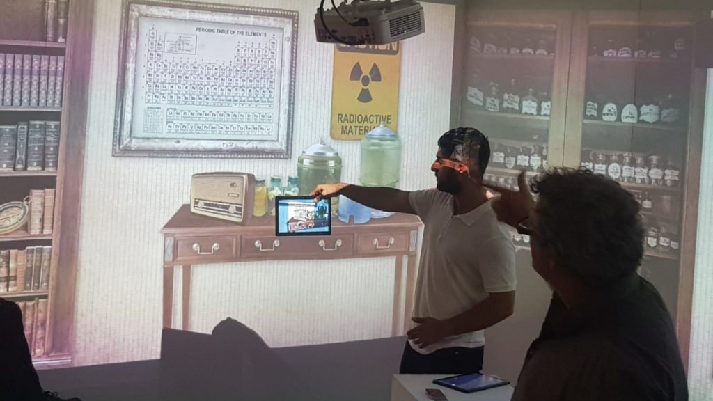 Photo of a man holding a tablet up in front of a projected Victorian laboratory scene — a periodic table, a radioactive-materials sign and shelves of glass bottles — while another participant watches, showing a tablet used alongside the wall projection