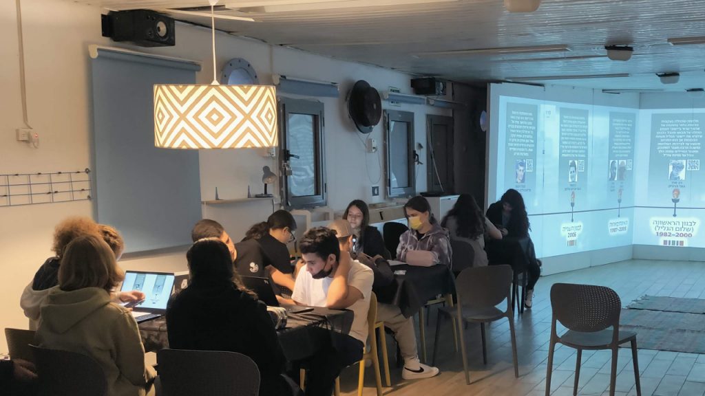 Photo of students working on laptops at tables while a Hebrew timeline of Israel's Lebanon campaigns, marked 1982 to 2000 and 1997 to 2003, is projected across the three walls behind them