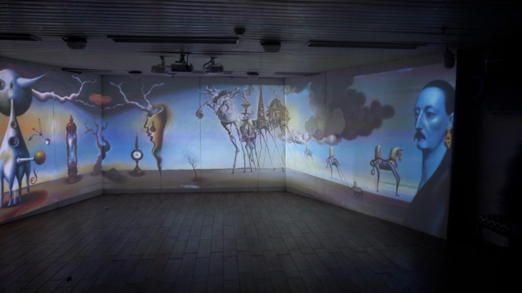 Photo of a three-wall immersive projection room showing a surreal Dalí-style panorama — melting clocks, spindly stick-legged creatures, and a man's head in profile — projected across the walls, with ceiling projectors and a wood-plank floor visible