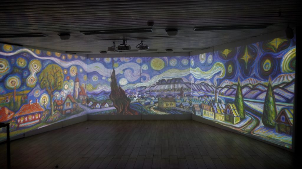 Photo of the same three-wall immersive room showing a Van Gogh "Starry Night"-style panorama — a swirling sky, a village with red-roofed houses, and a tall dark cypress tree — projected across the walls, with ceiling projectors visible