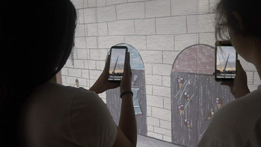 Photo taken over the shoulders of two students holding phones up to a drawn Jerusalem stone wall with arched doorways; on both screens an augmented-reality layer replaces the drawing with a photograph of the Chords Bridge, captioned with its Hebrew name