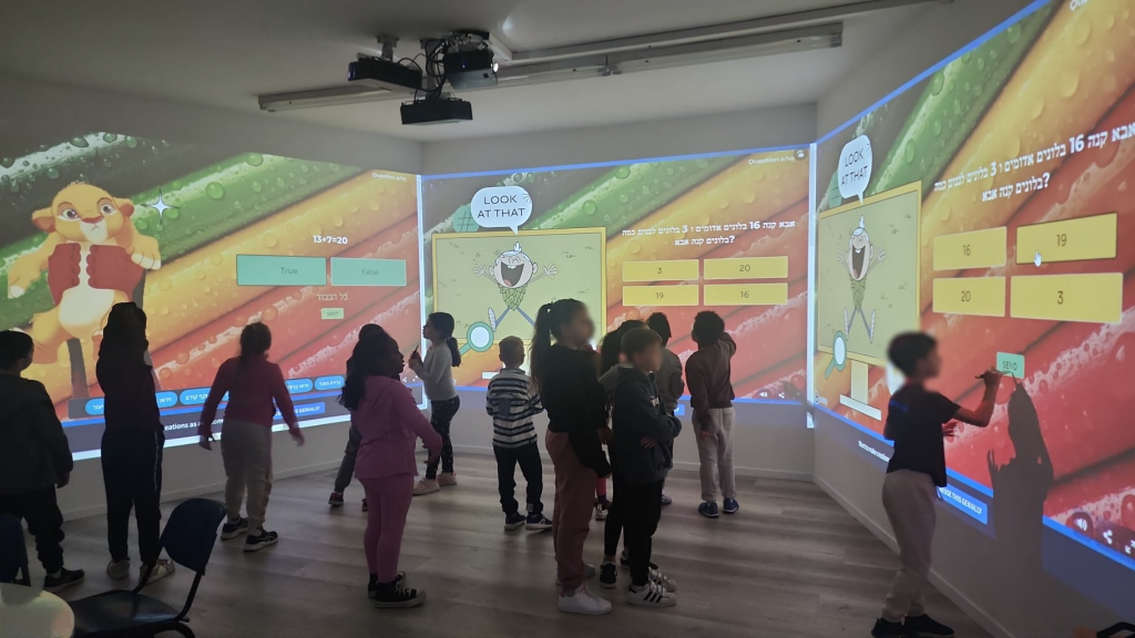Photo of a class standing in the immersive room during a maths activity: a Hebrew word problem about balloons with multiple-choice answer tiles spans the centre and right walls, an animated cub character fills the left wall, and one student reaches up to touch an answer