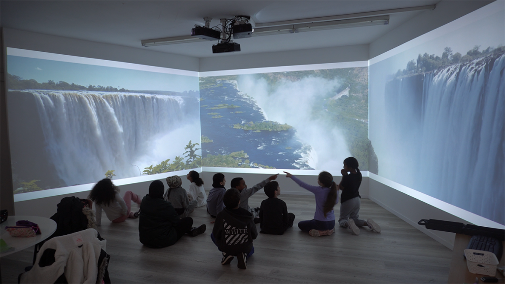 Photo of children sitting on the floor of the immersive room facing a panorama of Victoria Falls that runs continuously across all three walls, with one child pointing at the falling water