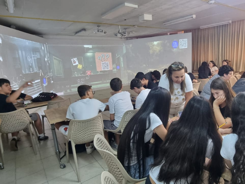 תצלום של פעילות קבוצתית בחדר אימרסיבי: תלמידים יושבים סביב שולחנות ומורה רכונה מעליהם ומשוחחת איתם, וברקע מוקרן על הקירות חדר בריחה ובו סימני שאלה וקוד QR