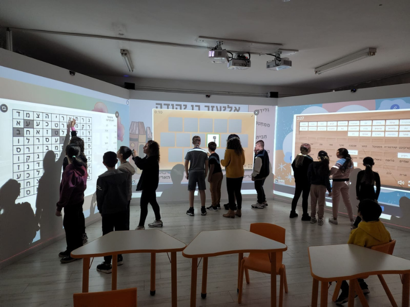 תצלום של כיתה בחדר אימרסיבי: כעשרה תלמידים עומדים מול שלושה קירות הקרנה – משמאל תפזורת אותיות בעברית, במרכז משחק התאמת כרטיסים בנושא אליעזר בן יהודה ומימין לוח מילים – עם מקרנים על התקרה ושולחנות וכיסאות כתומים בחזית