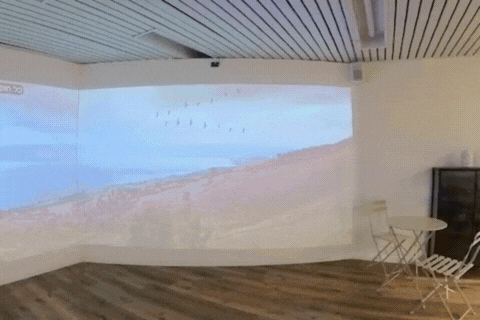 אותו חדר מעוקל, פעם עם הקרנת נוף חוף וגבעות על קירות המסך ופעם מואר כפינת ישיבה עם שולחן, כיסאות וספה
