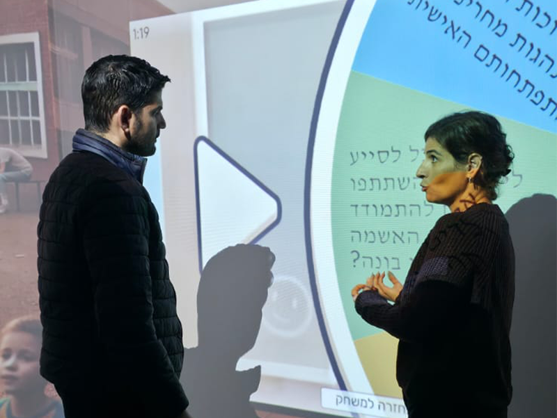 איש נוגע בקיר מוקרן עם כפתור הפעלה גדול, לצידו אישה מדברת ומחווה בידיה מול טקסט בעברית על המסך